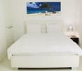 A modern white bedroom with minimalist design, featuring a large white bed, matching white pillows, a beach-themed painting, and a bedside table with a lamp.