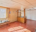 Spacious living room with wooden paneled walls, a tiled floor, and a large window. The room features a wooden cabinet with dishes and decorative items, as well as a green vintage-style refrigerator. There is also a table with tea cups and a serving tray,