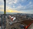 The image shows a construction site with various buildings and cranes in the background. The sky is filled with clouds and the sun is setting, creating a warm, golden glow. There are several construction vehicles and equipment visible on the site.