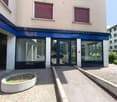 The image shows a commercial building with a blue awning and large glass windows. The building has a beige exterior and there is a raised concrete platform with a planter in front of the entrance. The surrounding area appears to be a residential neighborh