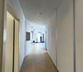 The image shows a long, well-lit hallway with a white ceiling and walls. The floor is made of hardwood or a similar material. There is a person visible in the distance, walking down the hallway. The hallway appears to be part of an office or commercial bu