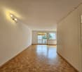 The image shows a spacious apartment with a large open floor plan. The room has white walls and a parquet wood floor. There are two windows that provide natural light and a view of the surrounding area. The room appears to be a living/dining area, with a