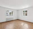Empty room with white walls, hardwood floors, and two large windows providing natural light. The room appears to be a living room or bedroom with a radiator heating system.