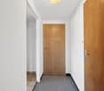 The image shows a hallway with white walls, a wooden door, and a tiled floor. The hallway appears to be simple and functional, with minimal decor.
