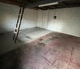 The image shows a small, dimly lit storage room with white brick walls and a concrete floor. There is a ladder or shelving unit visible in the corner, and the room appears to be empty and in need of some cleaning or renovation.