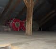 The image shows the interior of an attic space. The ceiling is made of wooden beams and the floor is covered with insulation material. A red sleeping bag or cushion is visible in the corner, suggesting this space may be used for storage or occasional slee