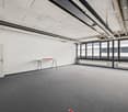 Large open office space with concrete ceiling, exposed ductwork, and gray carpeted floor. The room has large windows providing natural light and a small table near the entrance.