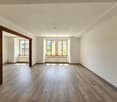 Spacious living room with hardwood flooring, large windows providing natural light, and wooden door frames.