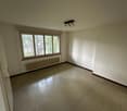 Empty room with white walls, hardwood floor, and a large window overlooking trees outside. The room has a ceiling light fixture and a radiator heater along the wall.
