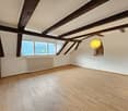 Empty room with wooden floor, white walls, wooden beams, window with mountain view, hanging lamp, and white radiator.