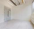 An empty room with tiled flooring, white walls, white door, and a radiator on the right side. There are windows and a white door, and a white ceiling with a light fixture