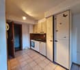 Small kitchen with white tiled floor, brown tiled wall, cabinet space, and built-in refrigerator. It has a gas oven, sink with faucet, and light in the ceiling.