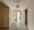 Long hallway with white tiled flooring, wooden cabinets, white walls, single hanging lamp, two doors on the right side, small balcony at the end