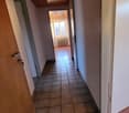 Tiled floor, ceiling with ceiling lights, walls are painted white, radiator on one wall, doorway in the middle, door on the left side.