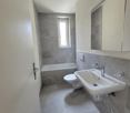 The image shows a modern bathroom with gray tile walls and floors, a white toilet, a white sink with a faucet, and a bathtub with a window.