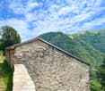 Old stone house with red roof, situated in a mountainous area with lush green vegetation