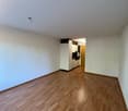 Empty room with wooden floor, white walls, a kitchen with cabinets, an open door to a hallway