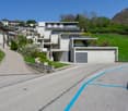 The image shows a residential area with modern multi-story apartment buildings nestled into a hillside. The buildings have a clean, minimalist design with large windows and balconies. There is a paved road with a blue line marking running through the scen