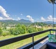 Balcony overlooking a scenic landscape with mountains, forests, and a village in the distance. The balcony has a black railing and a decorative tropical-themed pillow.