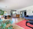 Spacious open-plan living area with hardwood floors, large windows, built-in bookshelves, potted plants, blue sofa, wooden dining table and chairs, and various artwork and decor on the walls.