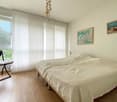 Spacious bedroom with large windows, white walls, hardwood flooring, a bed with a light-colored bedspread, and a framed artwork on the wall.