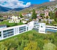 large white building with many windows and balconies, surrounded by green grass and trees, mountains in the background