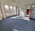 Large open office space with floor-to-ceiling windows, providing a bright and airy atmosphere. The room features a gray carpeted floor, white walls, and a suspended ceiling with air conditioning vents. The space appears to be suitable for use as an office