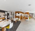 Large open-plan space with modern furniture and decor, including white tables and chairs, a curved wooden reception desk, and shelving units displaying various products. The space has a sleek and contemporary design with tile flooring and recessed lightin