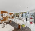 The image shows the interior of a modern retail store or showroom. The space features a clean, minimalist design with white walls, wood accents, and glass display cases. There are various products and displays showcased throughout the room, including what
