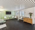 Modern office space with green walls, white chairs, glass tables, TV, and a wooden cabinet with a dishwasher visible.