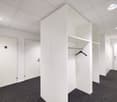 The image shows a modern office space with white walls, a carpeted floor, and recessed lighting. There are two doors visible, likely leading to other rooms or offices. In the center of the image, there is a white wardrobe or closet unit with a clothes han