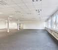 Large open office space with white walls, gray carpeted floor, suspended ceiling with recessed lighting, and large windows providing natural light. The space appears to be empty and ready for use.