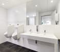 Large, modern bathroom with white tiled walls, black tile flooring, two white sinks with chrome faucets, and a dishwasher visible.