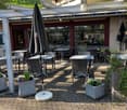 A restaurant's outdoor dining area with brick floors, round tables, and woven chairs. Potted plants and umbrellas add a touch of greenery and shade. Balconies and plants on the upper floors add to the charm. Behind the restaurant, a building's windows and