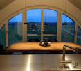 Dining area with long wooden table, 3 black chairs, metal balustrade, stainless steel sink, faucet, long windows with view to the landscape