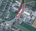 The area features a dense industrial setup with several large buildings, and various facilities, surrounded by fields. Parking lots are visible, with several vehicles. The central focus is an area marked in red, likely indicating a specific plot or point