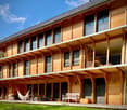 multi-story wooden house with balconies, large windows, outdoor furniture including bench and hammock, surrounded by grass