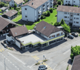 Aerial view of a neighborhood with several houses, some with brown roofs, some with white roofs, and some with balconies. The houses are surrounded by green grass and trees. There are cars parked in front of the houses and on the street.