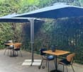 Outdoor seating area with tables and chairs under large umbrellas, surrounded by a hedge.