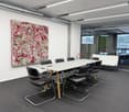 Modern office with a conference room setup. The conference room has a large white table surrounded by black chairs, a large colorful painting on the wall, and bright lighting from overhead lamps.