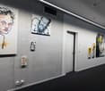 A long corridor featuring artwork on concrete walls. There are three larger paintings and three smaller paintings displayed. The flooring is black carpet.