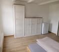 Empty room with wooden floor, white walls, white wardrobe, two large white cabinets, a white fridge, and a bed with white bedding.
