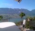 balcony with table and chairs, black railing, potted plants, lake and mountains in the background