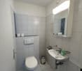 white tiled walls and floor, white sink, silver faucet, mirror with cabinet, toilet, trash bin, toilet paper