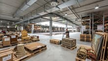large warehouse with many wooden pallets, boxes, and items, a person using a pallet jack, high ceilings, metal beams, and lighting