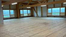 Large open space with wooden beams and trusses, hardwood flooring, and large windows providing natural light and a view of the surrounding landscape.