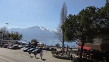 Outdoor parking lot with many cars, a lake, mountains, and a restaurant with outdoor seating.