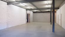 Large open space with white brick walls, a concrete floor, and a metal ceiling with exposed beams and lighting fixtures. The space appears to be a warehouse or industrial facility.