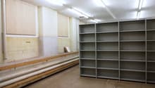 The image shows a large storage room with shelving units lining the walls. The room has a concrete floor and fluorescent lighting overhead. There are wooden benches or platforms along one wall, likely used for storage or work. The room appears to be a uti