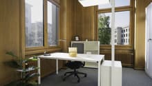A room with a desk, swivel chair, floor lamp, large windows, wooden walls, office storage, and a radiator.